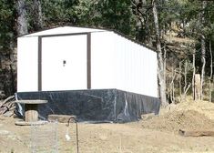 a small white shed sitting in the middle of a forest next to a pile of dirt