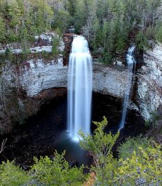 a large waterfall is coming out of a cave in the woods with trees around it