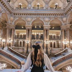 a woman with long hair standing in front of an ornate staircase and chandelier