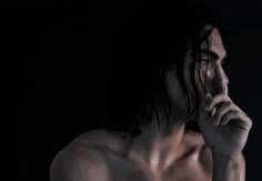 a man with long hair and no shirt holding his hand up to his face while standing in the dark