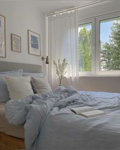 an unmade bed with blue sheets and pillows in front of two large windows that look out onto the trees outside