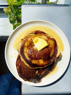 a stack of pancakes topped with butter and syrup on a white plate next to a potted plant