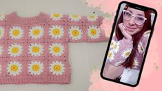 a pink crocheted sweater with white and yellow daisies on it next to an iphone