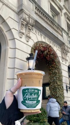 a cup of coffee is being held up in front of a building with christmas decorations