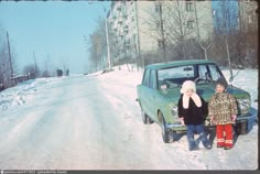 two children standing next to an old car on a snowy road in front of apartment buildings