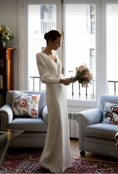 a woman standing in front of a window holding a bouquet of flowers and wearing a long white dress