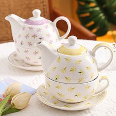 two teapots and saucers sitting on a table with flowers in the background