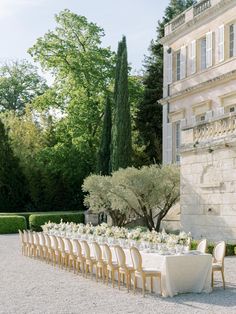 a long table set up with white linens and flowers in front of an old building