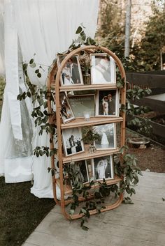 a wooden book shelf with pictures and greenery on the top, next to a white curtain