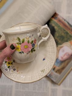 a person holding a cup and saucer with flowers on it in front of a magazine