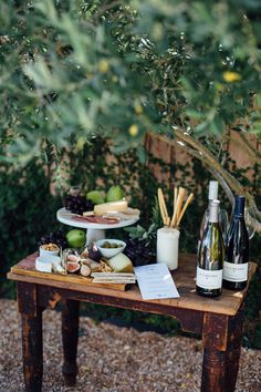 a wooden table topped with bottles of wine and cheeses next to a forest filled with trees