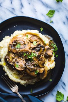 mashed potatoes topped with mushrooms and gravy on a black plate next to a fork