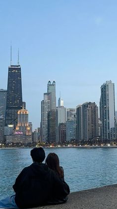 two people are sitting on the edge of a lake looking at the city