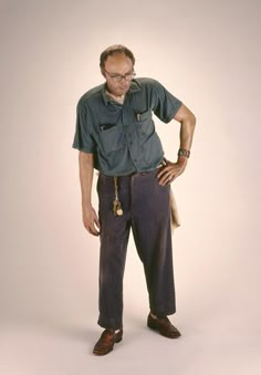 an older man with glasses standing in front of a white background wearing blue pants and a green shirt