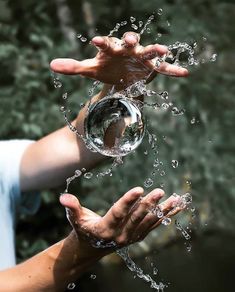 two hands are holding a glass ball with water splashing out of it to the camera