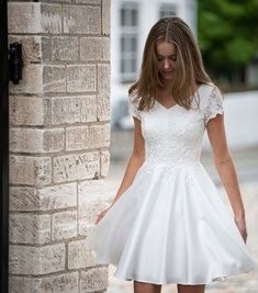 a woman in a short white dress is standing by a brick wall and looking down