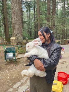 a woman holding a small white dog in her arms while standing next to a forest