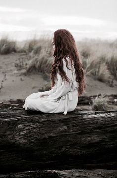 a woman with long red hair sitting on a log in front of some grass and sand