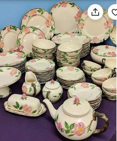 a table topped with lots of white dishes and cups filled with flowers on them next to a purple table cloth