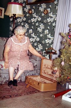 an old woman sitting on a couch next to a christmas tree with her suitcase open