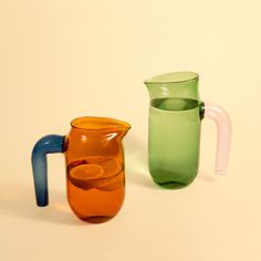 three different colored glass pitchers sitting next to each other on a white surface, one has a blue handle and the other has an orange handle