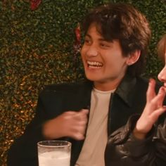 two people sitting next to each other in front of a wall with plants on it