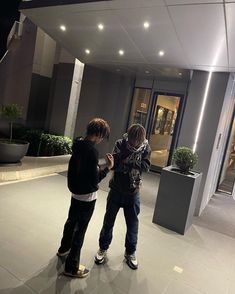 two young men standing in front of a building at night with their cell phones open