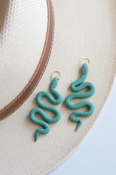 two green earrings sitting on top of a white hat next to a tan straw hat