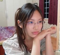 a young woman with glasses sitting in front of a mirror looking at the camera while holding her hand on her chin