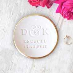 a wedding ring sits on top of a white plate next to pink flowers and a pair of earrings