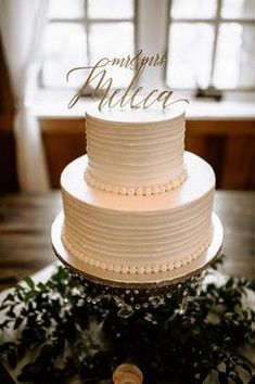 a white wedding cake sitting on top of a table next to a window with greenery