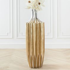 a gold vase with flowers in it sitting on a wooden floor next to a white wall