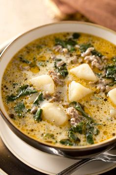 a bowl of soup with potatoes and spinach on a saucer next to a spoon