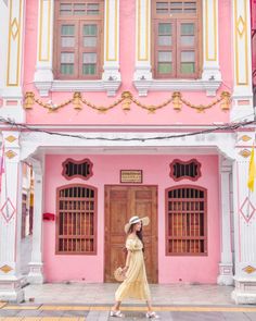 a woman standing in front of a pink and white building with wooden doors, wearing a straw hat