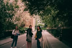 three women standing on the side of a road next to trees and bushes, with buildings in the background