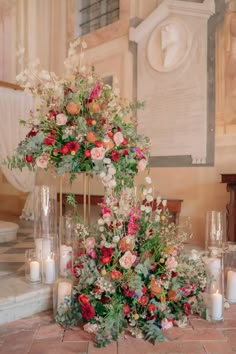 an arrangement of flowers and greenery on display in front of a wall with candles