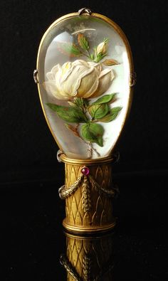 a white rose in a glass dome with gold trim on a black surface, surrounded by green leaves