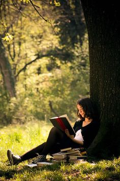 a woman sitting on the ground reading a book next to a tree in a forest