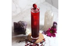 a tall glass filled with red liquid next to rocks and flowers on a marble surface