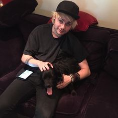 a person sitting on a couch with a dog in his lap and wearing a hat