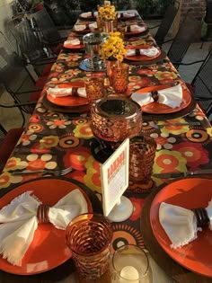 a long table with orange plates and place settings on it, along with flowers in vases