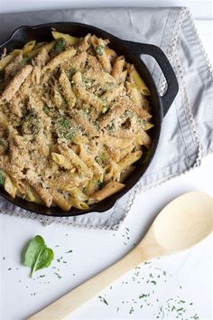 a skillet filled with pasta and pesto on top of a white table next to a wooden spoon