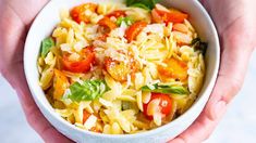 two hands holding a bowl of pasta with tomatoes and basil on top, while another hand holds the bowl