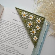 an open book with white flowers on it and a green triangle shaped brooch in the middle