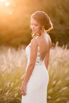 a woman in a white wedding dress standing in tall grass at sunset with her back to the camera