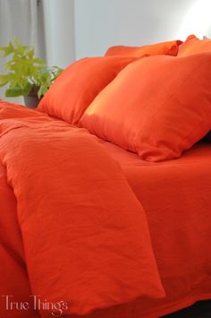 an orange comforter and pillows on a bed in a room with white walls, potted plant and window