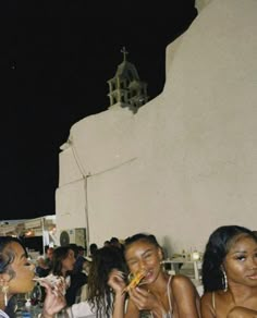 three women sitting at a table eating food and drinking wine in front of a white building