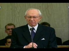 an older man in a suit and tie standing at a podium with other people behind him