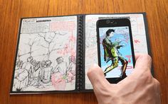 a hand holding an open cell phone in front of a book with drawings on it