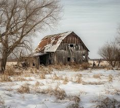 an old barn sits in the middle of a snow covered field with trees and bushes